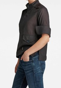 Personne portant une chemise boutonnée à manches courtes sombre avec une poche sur un jean en denim bleu, main partiellement dans la poche, sur fond blanc.