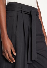 Gros plan sur une personne portant un pantalon noir taille haute avec une ceinture en tissu nouée à la taille et un top sans manches blanc.