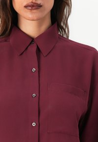 Femme portant une chemise rouge foncé boutonnée avec un col pointu et une poche poitrine, lèvres avec un rouge à lèvres brillant foncé et un grain de beauté au-dessus de la lèvre supérieure.