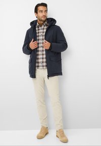 Uomo che indossa un giubbotto invernale blu con cappuccio sopra una camicia a scacchi marrone, pantaloni beige e sneakers color sabbia, in piedi su uno sfondo bianco.