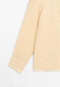 Beiges langärmeliges Strickoberteil mit Bündchenärmel vor einem hellgrauen Hintergrund.