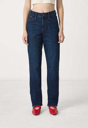 Vrouw draagt donkerblauwe jeans met rechte pijpen, cropped wit topje en rode balletpantoffels, staand tegen een effen achtergrond.