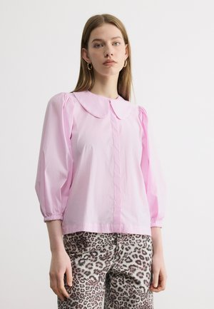 Žena s rovnými svetlohnedými vlasmi má zlaté kruhové náušnice, svetloružovú blúzku s nafúknutými rukávmi a golierom a nohavice s leopardím vzorom.