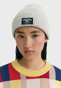 Ung person iført en cremefarvet strikket hue med "THINKING MU" label og en farverig ternet sweater med gul krave, neutral baggrund.