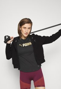Felpa nera con zip sopra una maglietta atletica grigia con logo "PUMA", abbinata a pantaloni corti compressivi borgogna con accenti e pattern arancioni.
