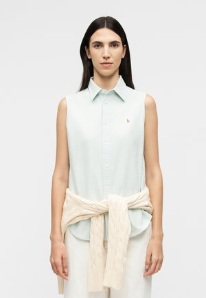 Γυναίκα με μακριά σκουρόχρωμα μαλλιά φοράει αμάνικο ανοιχτό γαλάζιο πουκάμισο με κουμπιά και κρεμ πουλόβερ με ανάγλυφα σχέδια δεμένο στη μέση, στέκεται μπροστά σε λευκό φόντο.