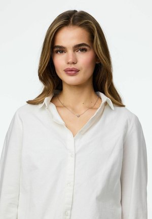 Biała koszula na guziki z luźnym kołnierzem, noszona na srebrnym naszyjniku z małym dekoracyjnym wisiorkiem. Miękka tekstura tkaniny, casualowy krój.