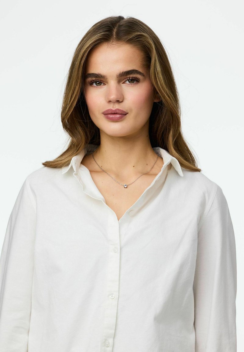 Chemise blanche à boutons avec un col décontracté, portée par-dessus un collier argenté orné d'un petit pendentif décoratif. Texture de tissu douce, coupe décontractée.