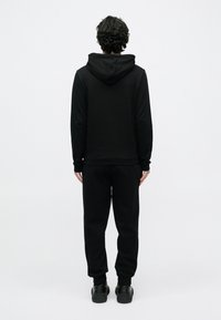 Fekete kapucnis pulóver, hozzáillő fekete edzőnadrággal párosítva. Mindkettő sima textúrájú, bordázott mandzsettákkal a bokánál és csuklónál.