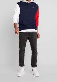Sudadera bloqueada en color azul marino, gris, rojo y blanco con un bolsillo frontal. Jeans negros y zapatillas blancas completan el atuendo.