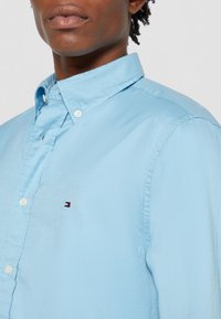 Camicia button-up azzurra chiara realizzata in tessuto liscio, con colletto, bottoni bianchi e un piccolo logo ricamato sulla zona sinistra del petto.
