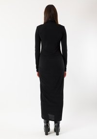 Abito nero a maniche lunghe in tessuto elasticizzato, con scollo alto, silhouette attillata e orlo lungo e fluente.