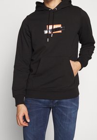 Sudadera negra con capucha, bolsillo canguro frontal y logo gráfico en rojo y blanco. Cuenta con cordones ajustables y puños acanalados.