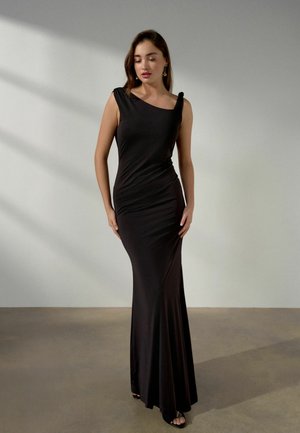 Frau in einem bodenlangen schwarzen Kleid mit Ein-Schulter-Trägern, stehend auf einem schlichten Boden vor einer hellgrauen Wand.