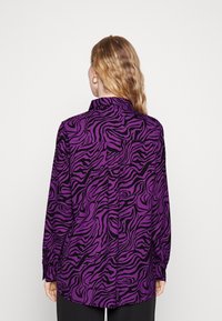 Femme aux cheveux blonds ondulés portant un haut à manches longues à rayures zèbre noir et violet, se tenant de dos contre un fond uni.