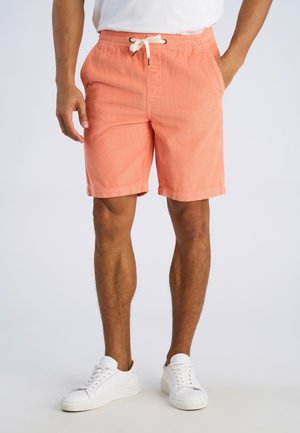 Mężczyzna w koralowych sztruksowych szortach z białym sznurkiem, białych trampkach i białej koszulce, stojący z rękami w kieszeniach.