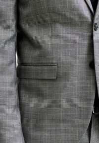 Gros plan d'une veste de costume grise à carreaux avec une poche à rabat et un bouton noir sur le devant, montrant la texture et le motif détaillés du tissu.