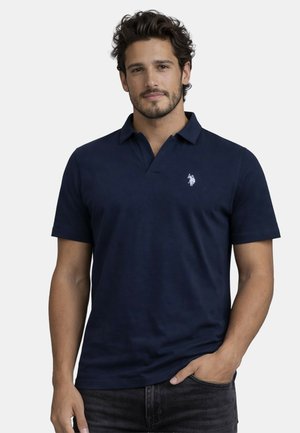 Junger Mann mit dunklem lockigem Haar, der ein marineblaues Polohemd mit kurzen Ärmeln und einem kleinen weißen gestickten Logo auf der Brust trägt.