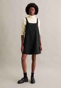 Zwarte denim mouwloze jurk met een vierkante halslijn, voorzien van een voorzak. Gepaard met een lichtgele coltrui en zwarte instappers.