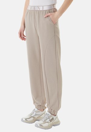 Pantaloni jogger beige con vita e polsini elasticizzati, mano in tasca, abbinati a sneakers bianche e oro con marchio "EA7".