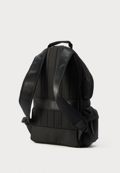 Μαύρο μαξιλαρωμένο σακίδιο με ρυθμιζόμενους ιμάντες, λαβή στο επάνω μέρος και μια μικρή πλευρική τσέπη με φερμουάρ, σε ουδέτερο φόντο.