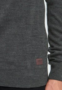 Dunkelgrauer Strickpullover mit gerippten Bündchen und Saum, aus strukturiertem Stoff und einem kleinen braunen Leder-Label am unteren linken Rand.