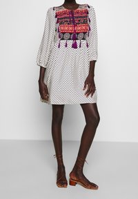 Kvinna klädd i en vit klänning med svarta mönster, röd och lila broderad krage, stående i bruna sandaler med blockklack och remmar.