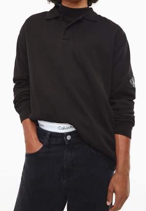 Zwarte langemouwen polo met een structuurstof en een kleine kraag, gecombineerd met donkere jeans met een zichtbaar taillebandlogo.