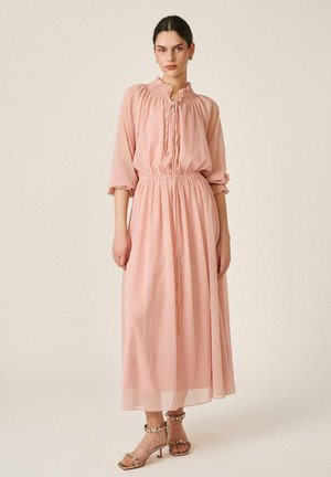 Mujer vistiendo un vestido largo hasta el tobillo, de manga larga y color rosa claro, con cuello con volantes y escote con lazo, combinado con tacones adornados con detalles dorados.