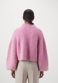 Kvinna med mörkt hår uppsatt i en tofs, iförd en strukturädd, överdimensionerad rosa tröja och ljusbeiga byxor, sedd bakifrån mot en enkel bakgrund.