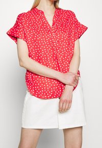 Czerwona bluzka z krótkim rękawem w białe kropki, z kołnierzykiem i zapinana na guziki, w zestawie z białą spódnicą. Gładka faktura materiału.