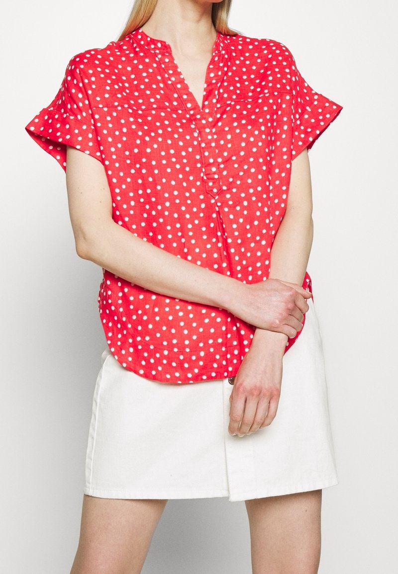 Czerwona bluzka z krótkim rękawem w białe kropki, z kołnierzykiem i zapinana na guziki, w zestawie z białą spódnicą. Gładka faktura materiału.