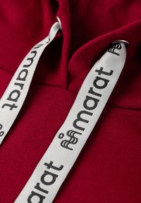 Close-up van rode hoodie-stof met witte trekkoorden door metalen oogjes, met zwarte "marat" tekst en logo.