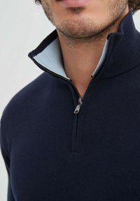 Pull en maille bleu marine avec un col zippé mi-hauteur ; présente une doublure intérieure bleu clair et des fermetures éclair en argent. Texture douce, design ajusté.