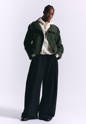 Green cropped jacket over a cream hoodie, paired with wide black trousers. Smooth fabric with layered design and tonal hardware accents.