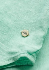 Muntgroene linnen stof met een gladde textuur, voorzien van een ronde gouden metalen tag met "Superdry" geschreven in donkere letters.