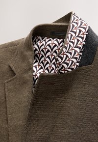 Blazer marron texturé avec une doublure de col à motif géométrique blanc, noir et marron et un patch intérieur sombre à l'épaule.