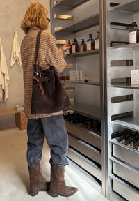 Brązowa torba na ramię ze skóry zamszowej z breloczkiem, koszula w kratę, wyblakłe dżinsy i brązowe kowbojki. Półki z różnymi butelkami i pudełkami.