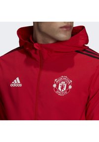 Chaqueta deportiva roja con capucha, detalles en negro y logo blanco del Manchester United en el pecho. Fabricada con un material suave y ligero.