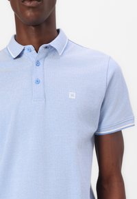 Heleheles sinise värviga polo särk tekstuurse kangaga, millel on kolme nupuga riba, kontrastne valge krae ja varrukate äär, ning väike logo.