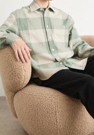 Personne portant une chemise à carreaux vert clair et beige assise sur un fauteuil beige texturé avec un pantalon noir.