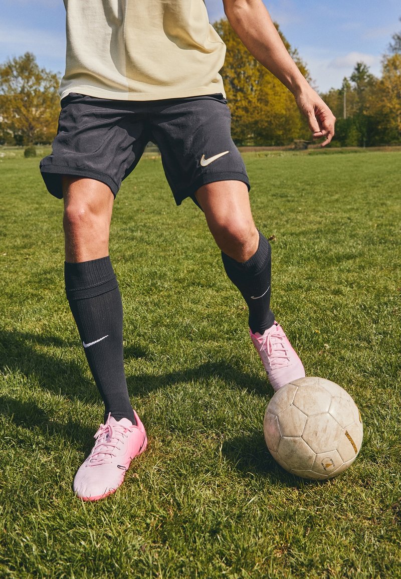 Shorts Nike noirs avec un logo doré, chaussettes noires jusqu'au genou et chaussures de football rose clair ; ballon de football sur un terrain de gazon.