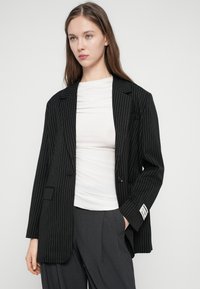 Čierne sako s tenkými prúžkami s výstrihom s výrezom, zapínaním na jeden gombík a náprsným vreckom, doplnené priliehavým bielym topom a sivými nohavicami.