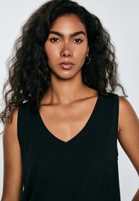 Joven con cabello rizado y oscuro que lleva una blusa negra sin mangas con escote en V, mirando directamente a la cámara con expresión neutra.