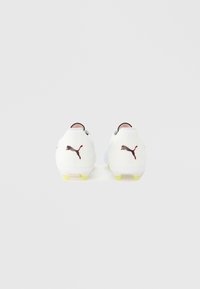 Białe korki piłkarskie z czarno-pomarańczowym logo Puma, żółtymi kolcami zapewniającymi przyczepność i eleganckim, nowoczesnym designem. Gładki materiał o minimalistycznym profilu.