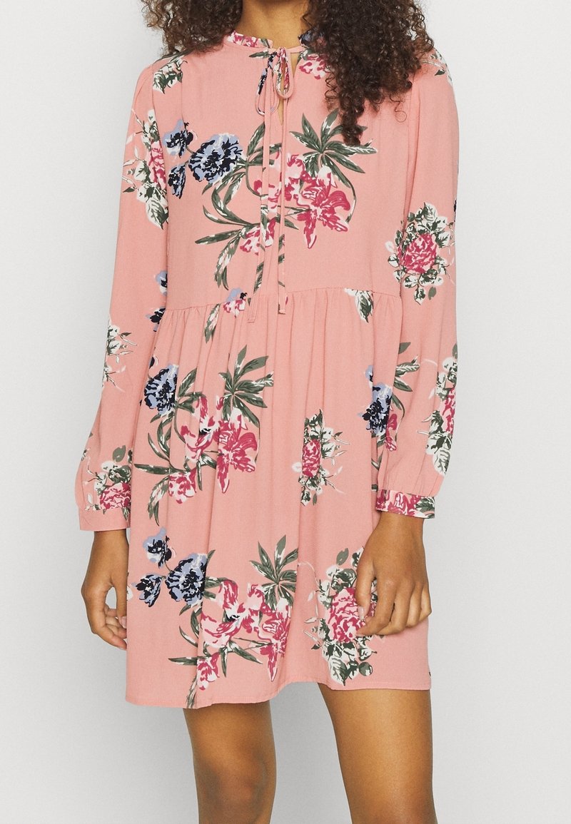 Vaaleanpunainen kukkamekko, jossa on pitkät hihat, nyöritys kaula-aukossa ja kerätty vyötärö. Sisältää sinisiä, punaisia ja vihreitä kukkakuoseja. Pehmeä kankaan rakenne.