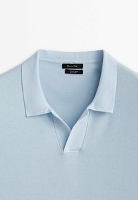 Polo en maille bleu clair avec un col doux et un détail d'encolure en V ouverte, montrant une étiquette de marque noire à l'intérieur du col.