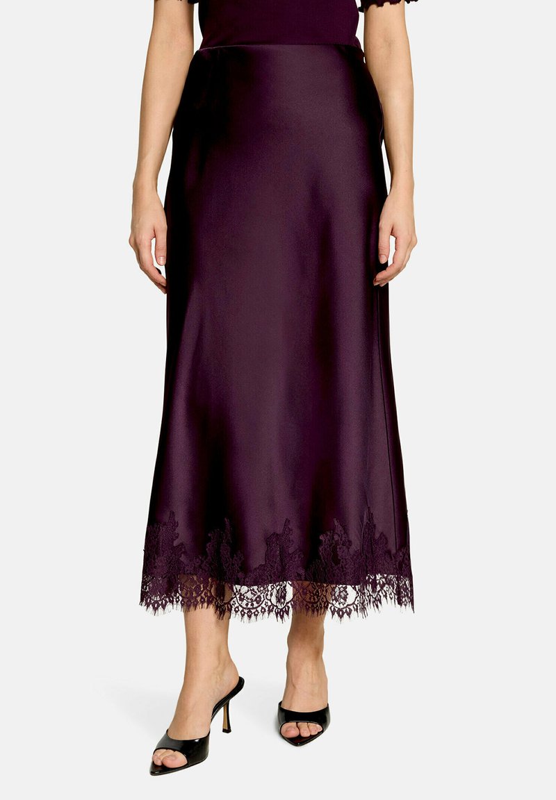 Femme portant une jupe midi en satin violet foncé avec un bord en dentelle à l'ourlet, associée à des chaussures à talons hauts noires ouvertes à l'avant.