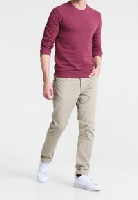 Man som går klädd i en långärmad vinröd tröja, beige byxor och vita sneakers med ena handen i fickan.