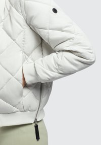 White quilted jacket featuring a zippered side pocket, ribbed cuffs, and a black button detail on the shoulder. Smooth texture and modern design.